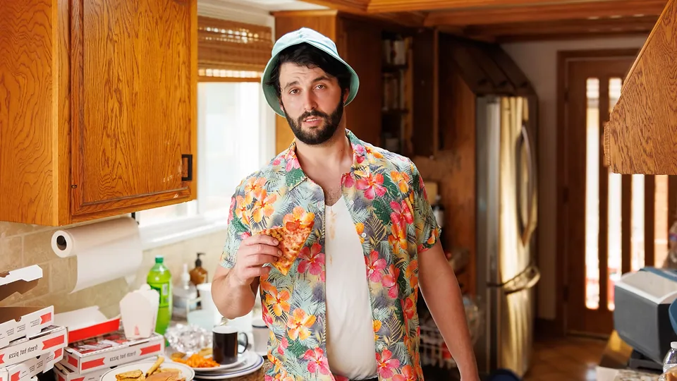 A man in a floral shirt holding a slice of pizza in a messy kitchen